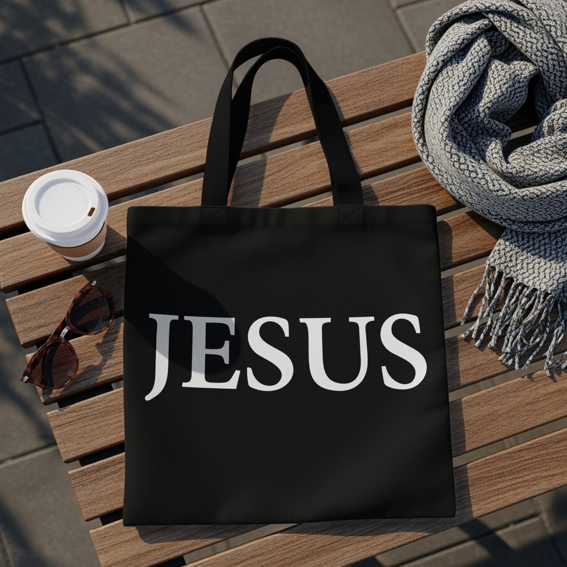 Black tote bag with "JESUS" printed in white on a wooden bench.