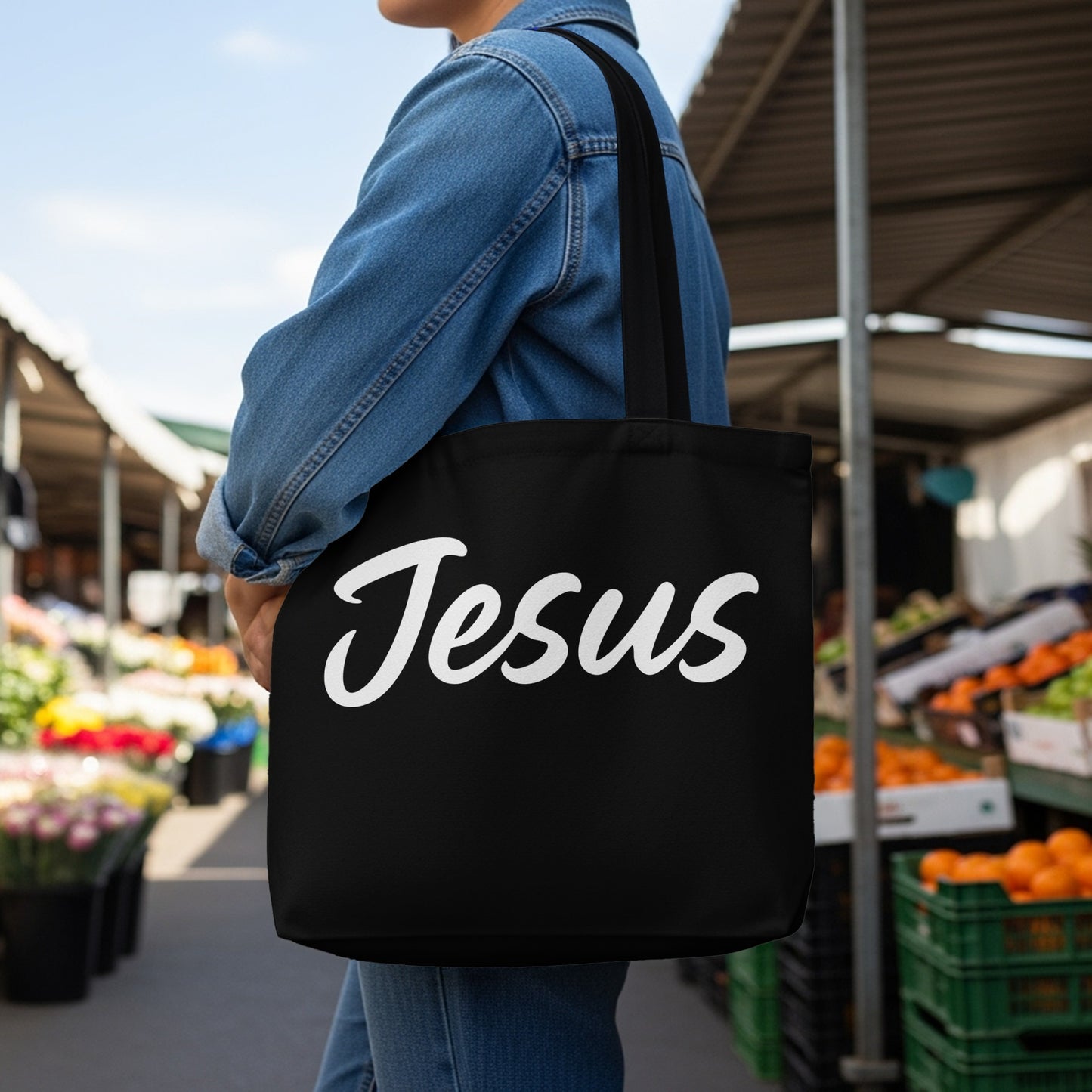 Black tote bag with "Jesus" written in white script held by a person in a denim jacket.