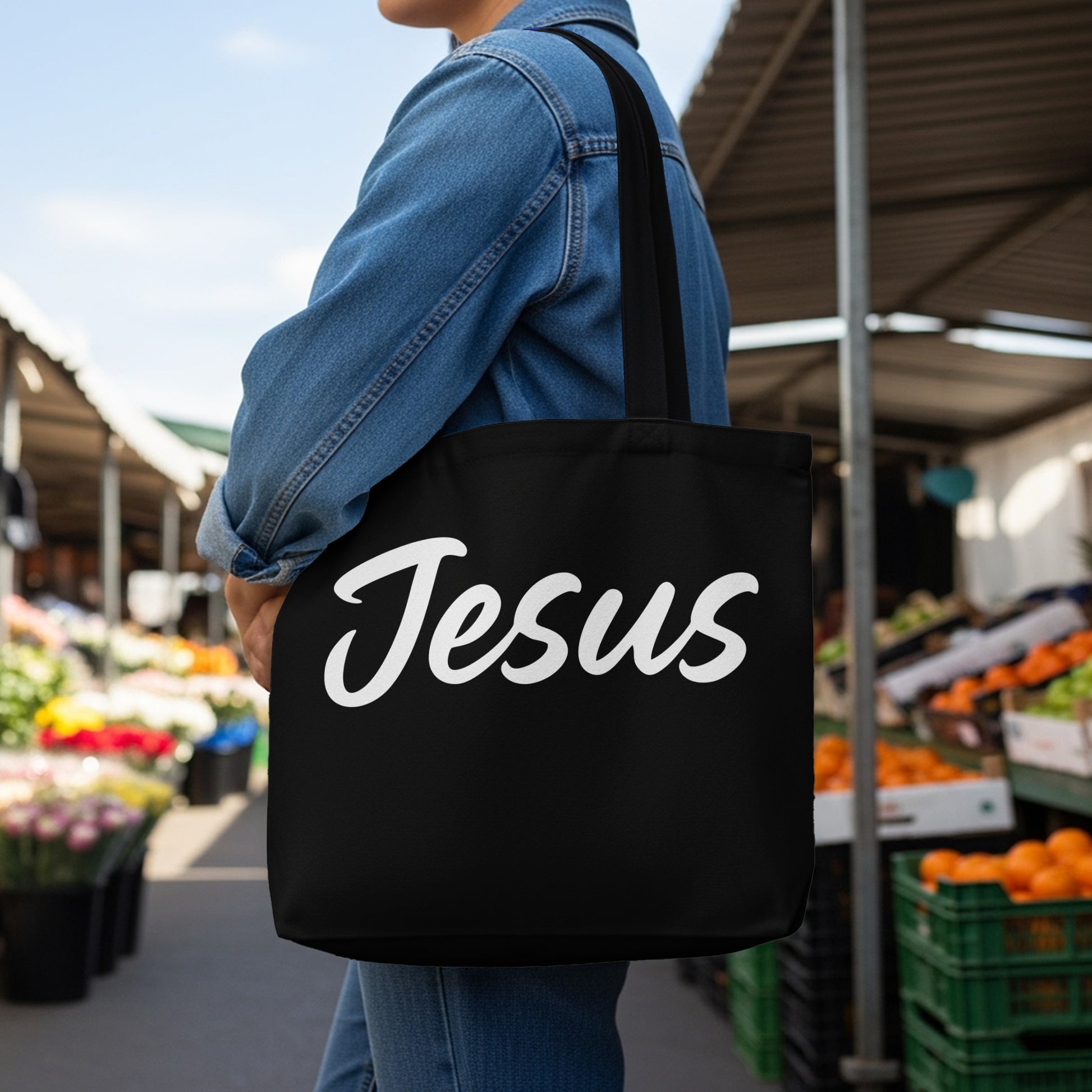 Black tote bag with "Jesus" written in white script held by a person in a denim jacket.