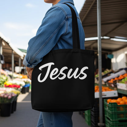 Black tote bag with "Jesus" written in white script held by a person in a denim jacket.