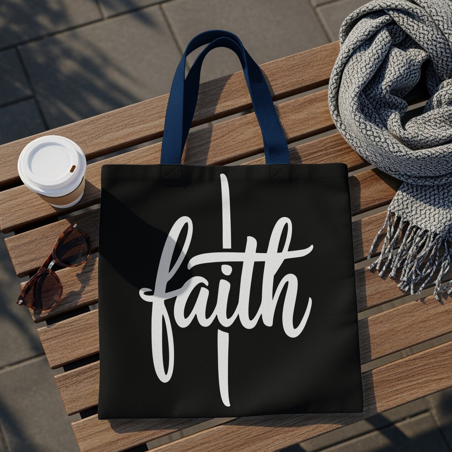 Black tote bag with "faith" in white script, on a wooden bench with a scarf and coffee cup.