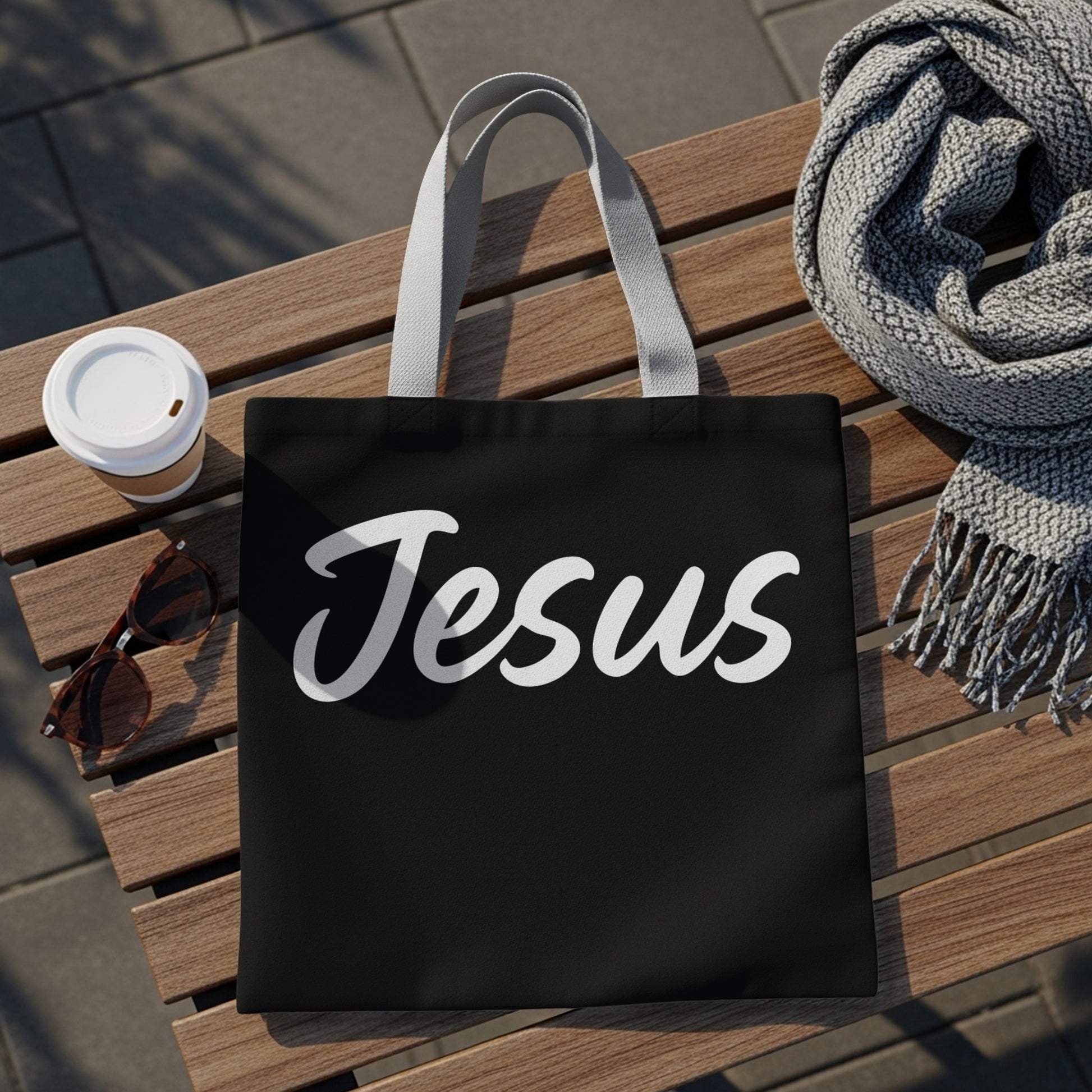 Black tote bag with "Jesus" in white script, next to coffee and sunglasses.