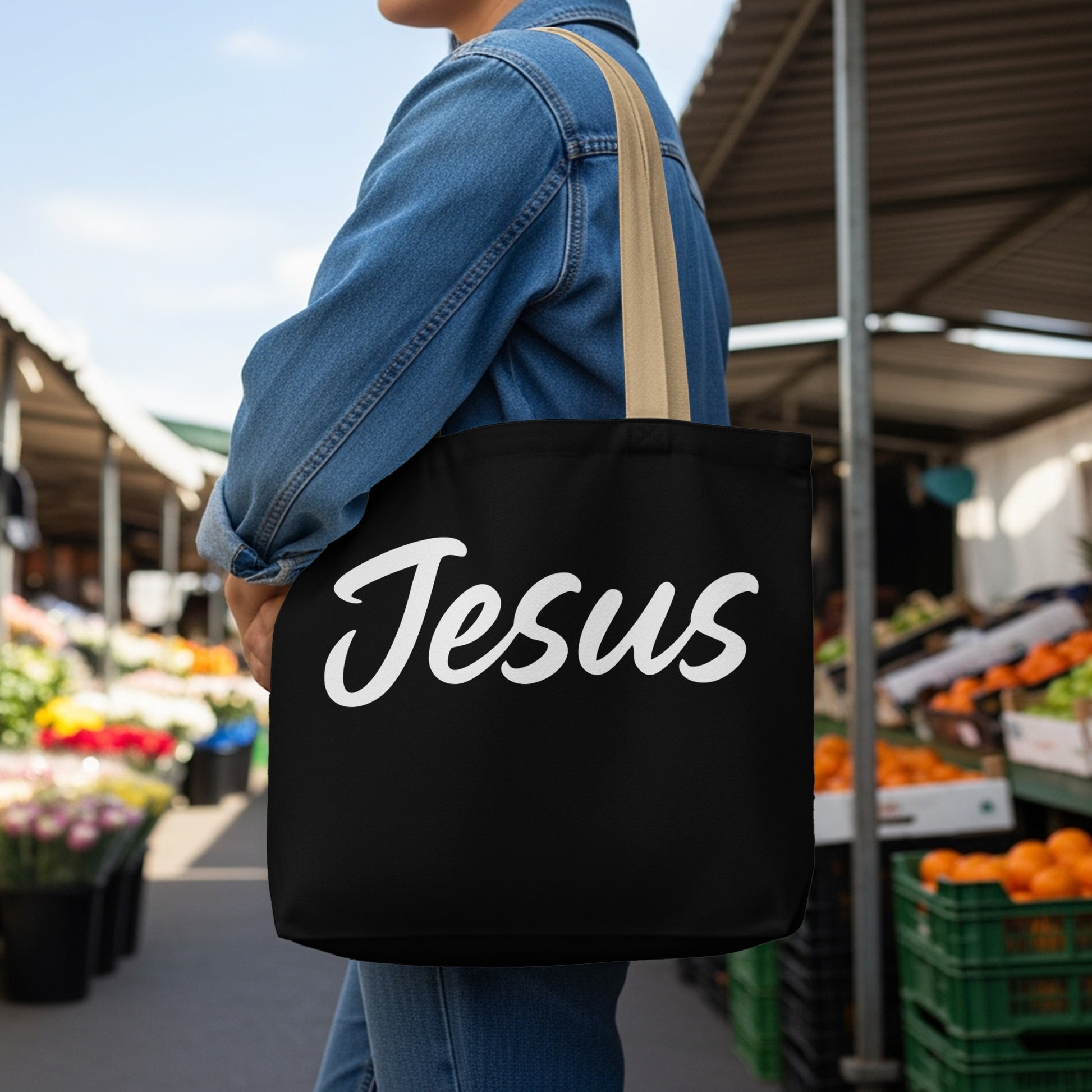 Black tote bag with white cursive text "Jesus" worn by a person outdoors.