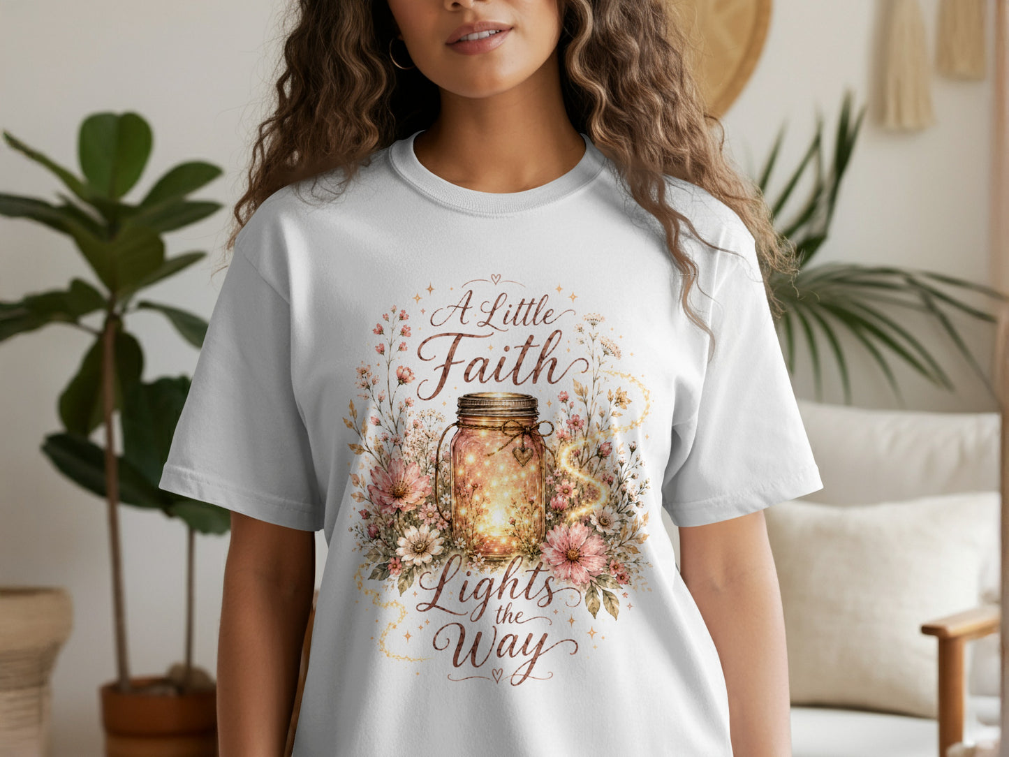 White t-shirt with a graphic featuring a glowing jar surrounded by flowers and the text "A Little Faith Lights the Way."