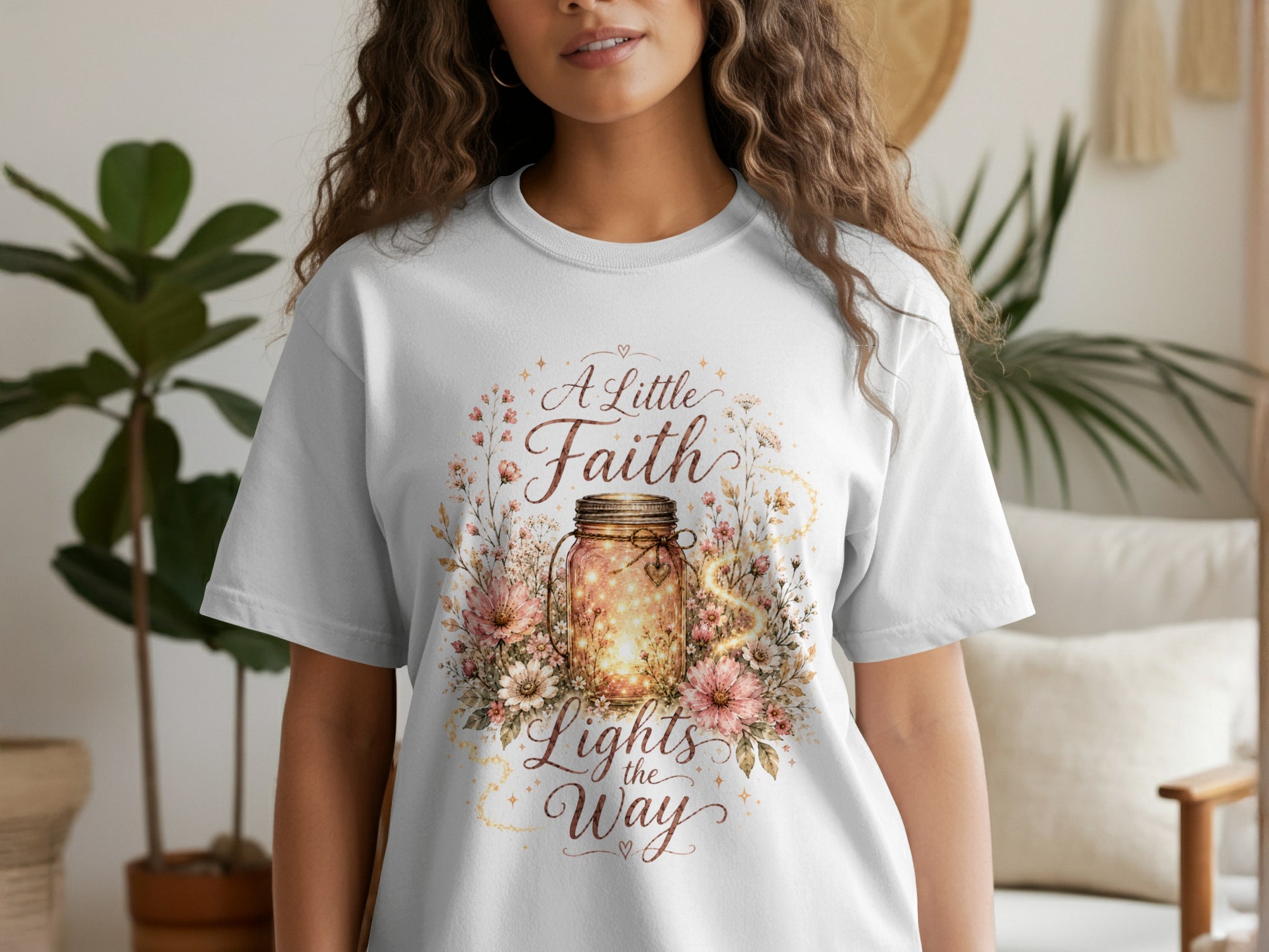 White t-shirt with a graphic featuring a glowing jar surrounded by flowers and the text "A Little Faith Lights the Way."