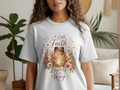 White t-shirt with a graphic featuring a glowing jar surrounded by flowers and the text "A Little Faith Lights the Way."