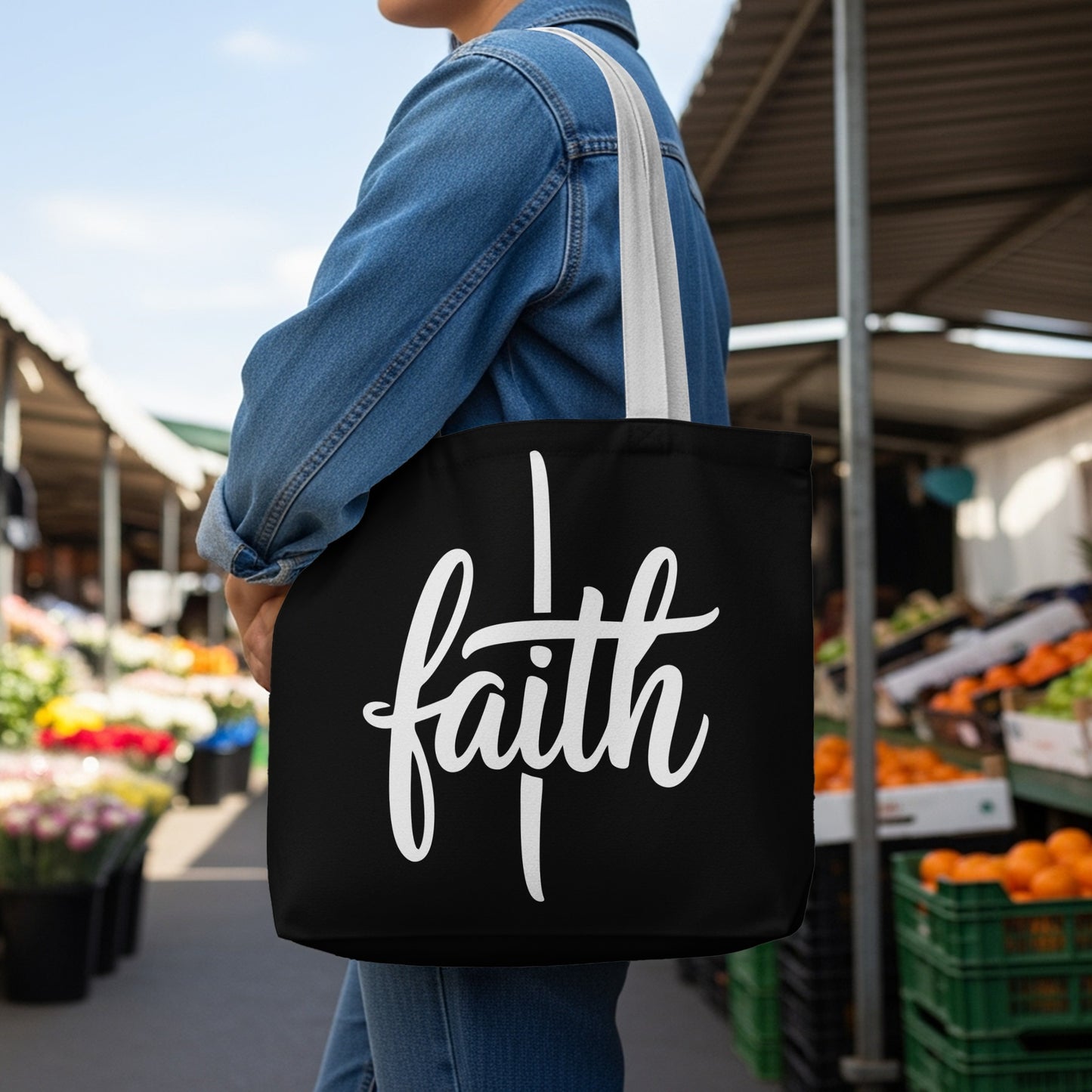 Black tote bag with white "faith" text and a cross symbol, worn by person in denim jacket.
