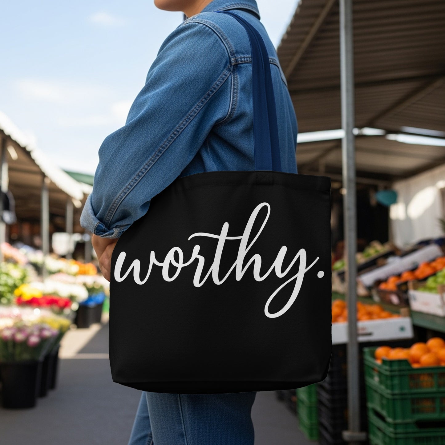 Black tote bag with white script text "worthy." against a market backdrop.