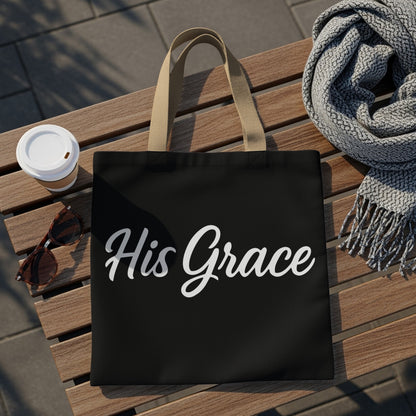 Black tote bag with "His Grace" text and tan straps, set on a bench.