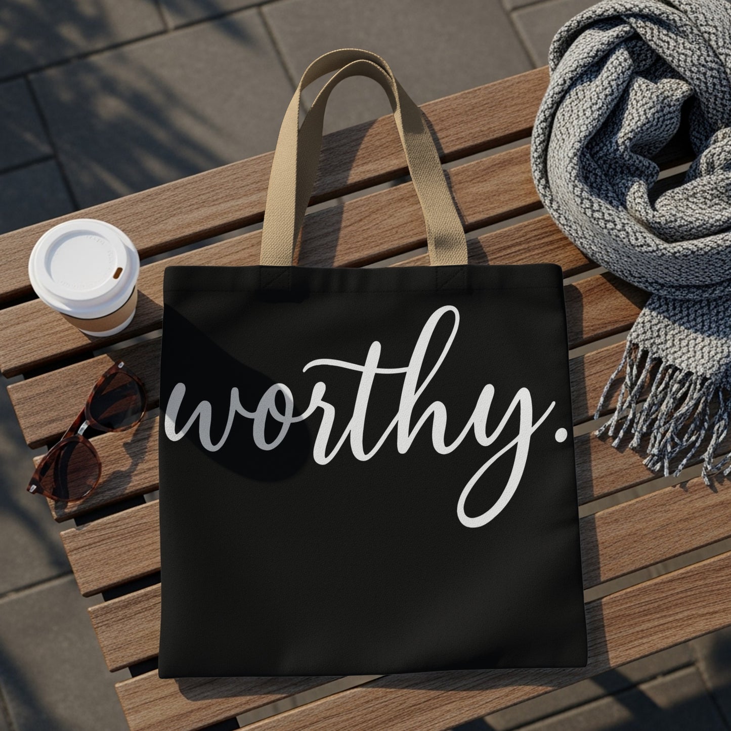 Black tote bag with white script text "worthy." beside coffee, sunglasses, and scarf.