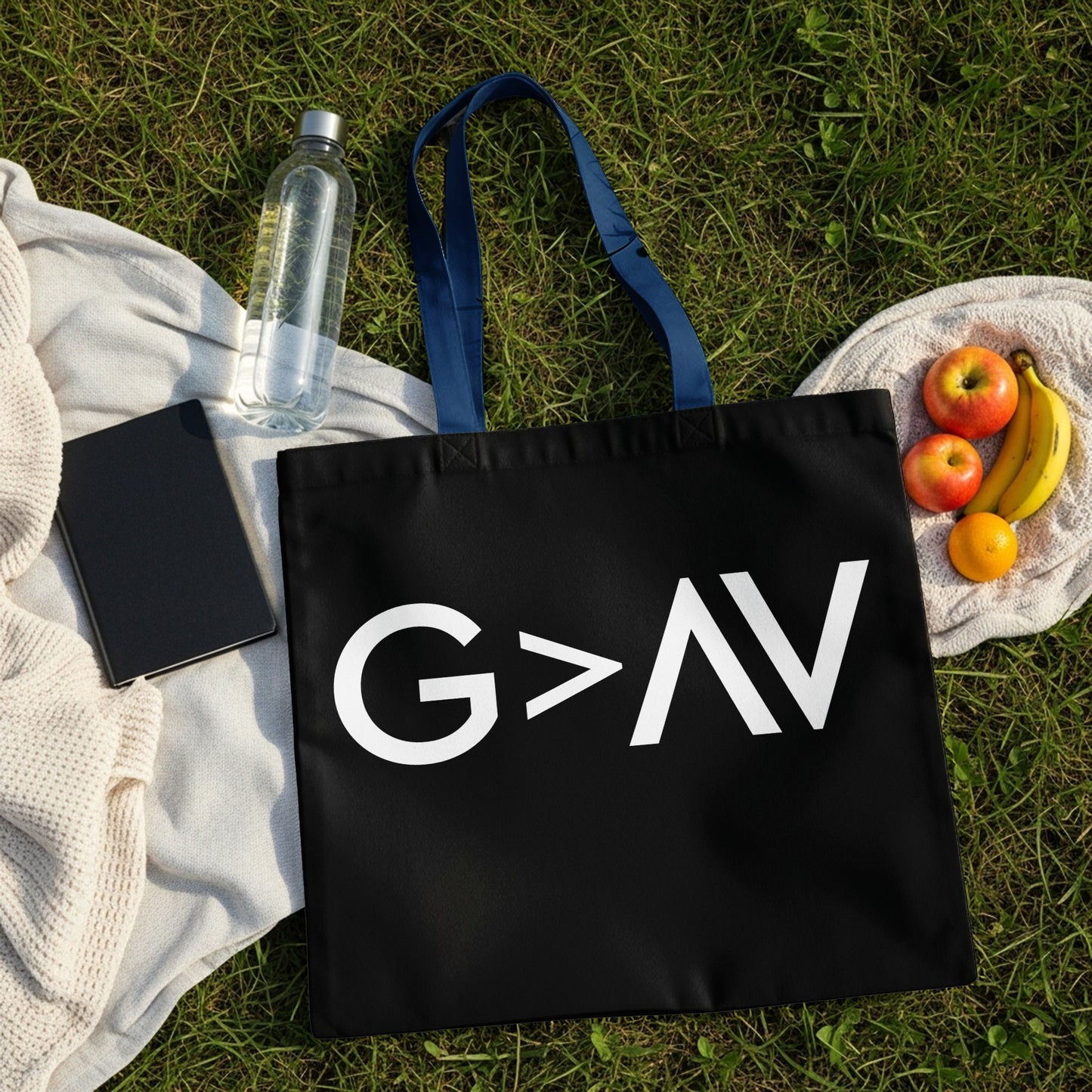 Black tote bag with white text "G>AV" on grass with fruit and water bottle.