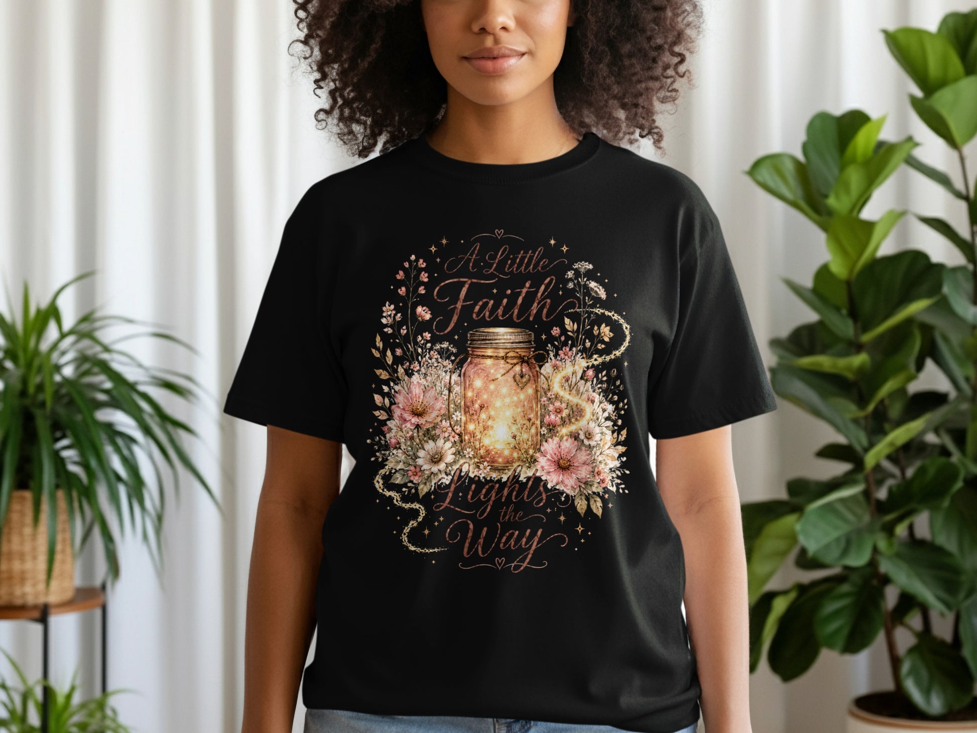 Black t-shirt with a floral design and a mason jar filled with lights. Text reads "A Little Faith Lights the Way".