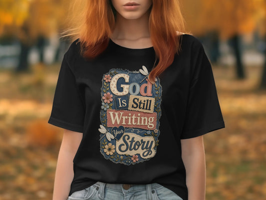 Black t-shirt with "God Is Still Writing Your Story" text and floral accents.
