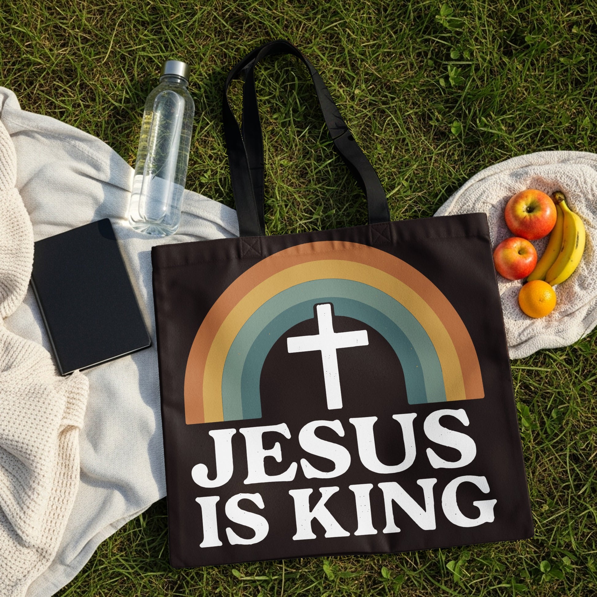 Black tote bag with rainbow, cross, and "JESUS IS KING" text on grass.