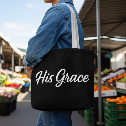 Black tote bag with white script text "His Grace" carried by a person in a denim jacket.