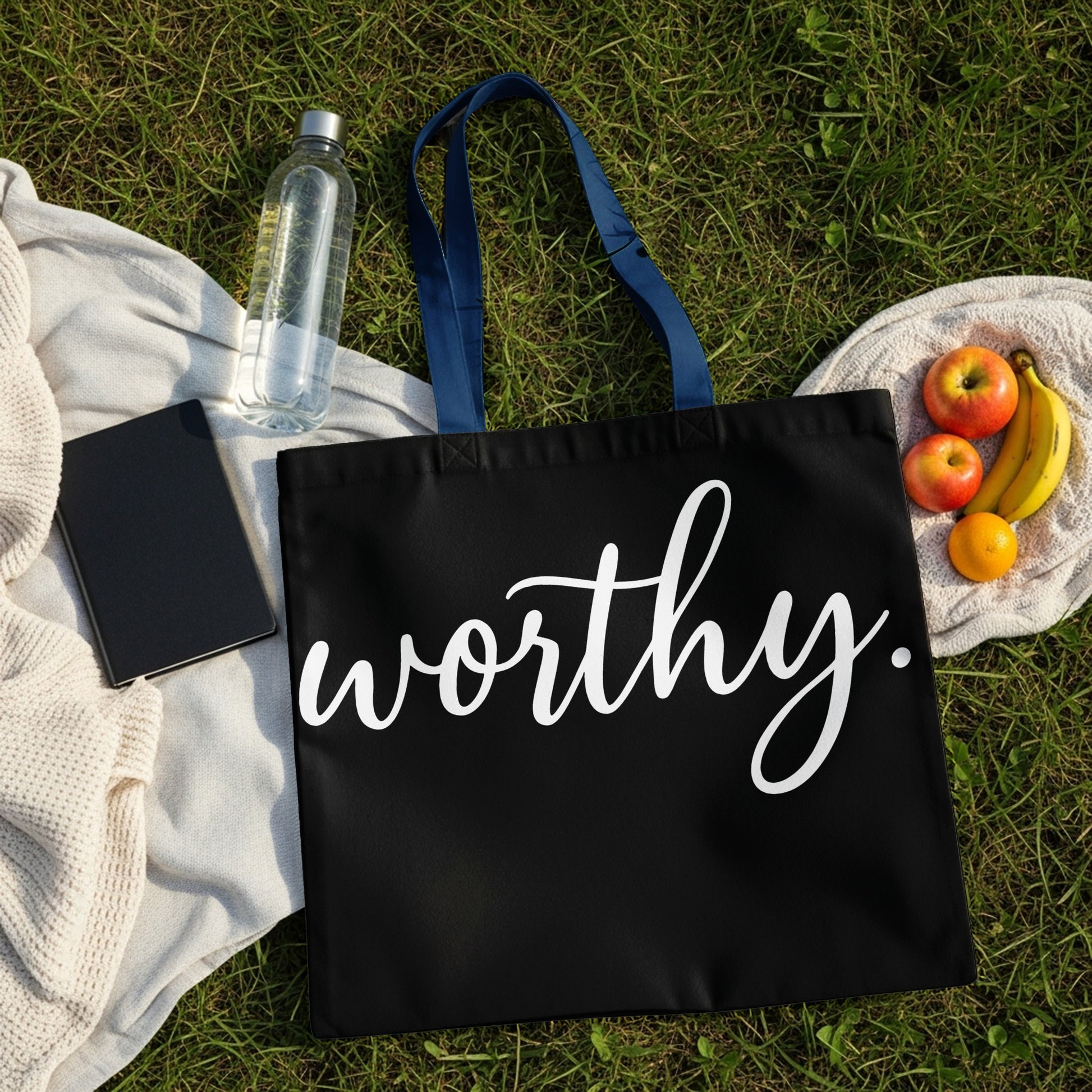 Black tote bag with white text "worthy." on grass with fruit and water bottle.