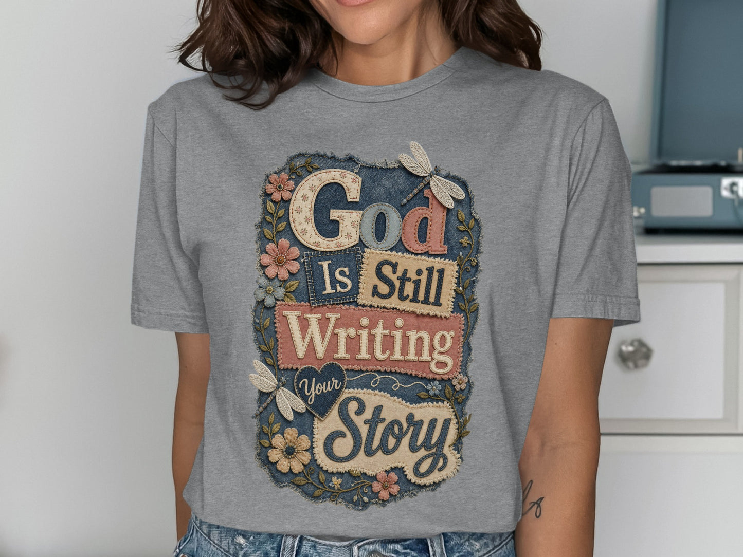 Grey t-shirt with "God Is Still Writing Your Story" text and floral dragonfly design.