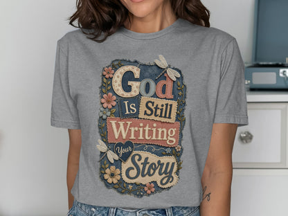 Grey t-shirt with "God Is Still Writing Your Story" text and floral dragonfly design.