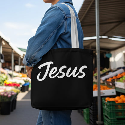 Black tote bag with "Jesus" in white cursive lettering, worn by a person outdoors.