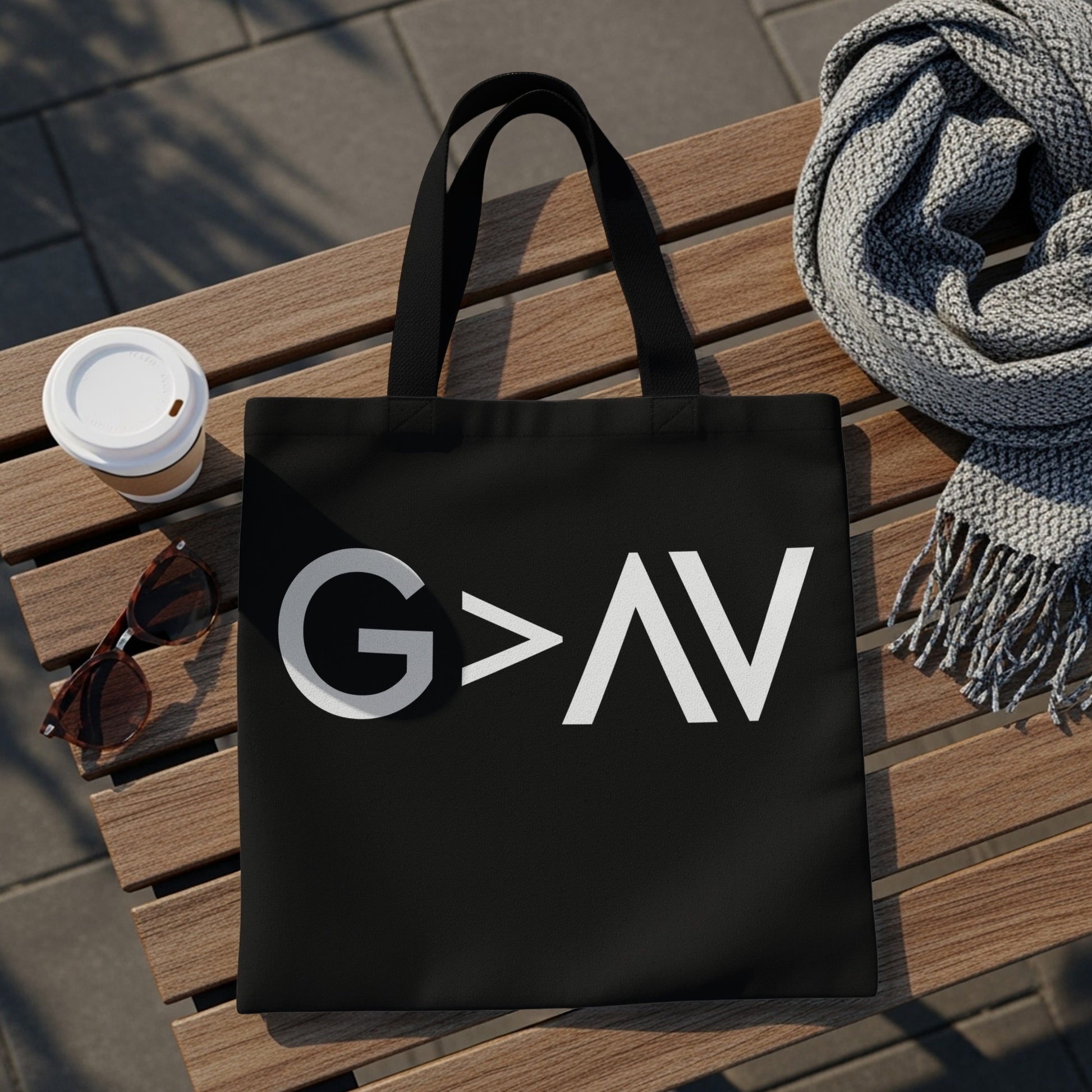 Black tote bag with white "G>AV" text, sunglasses, coffee, and scarf on bench.