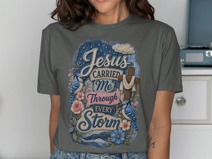 Jesus carried me through every storm text on a dark gray t-shirt with bluebirds and flowers.
