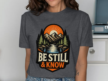 T-shirt graphic with mountains, lake, sun, trees, and text "Be Still & Know".