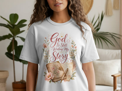 White t-shirt with "God Is Still Writing My Story" text, floral wreath, and open book.