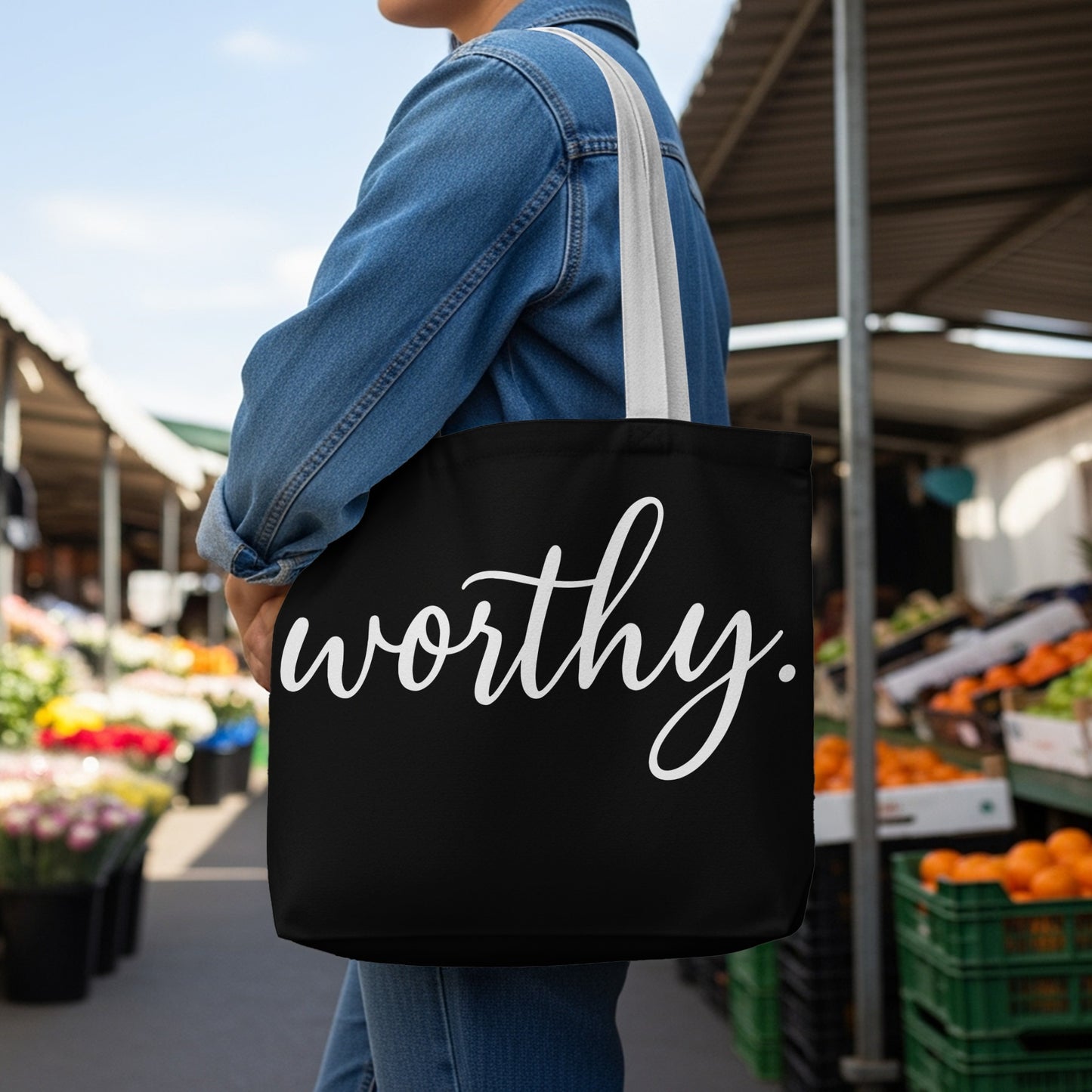 Black tote bag with white "worthy." text in script font worn by a person at an outdoor market.