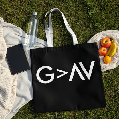 Black tote bag with white "G > AV" graphic, laid on grass with blanket, water bottle, notebook, and fruit.