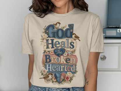 T-shirt with "God Heals The Broken Hearted" in textured letters and floral accents.
