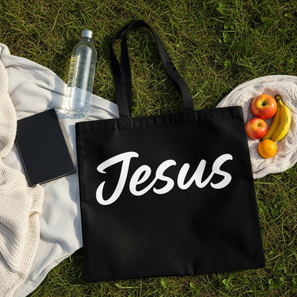 Black tote bag with white "Jesus" text, on grass with fruit and water bottle.