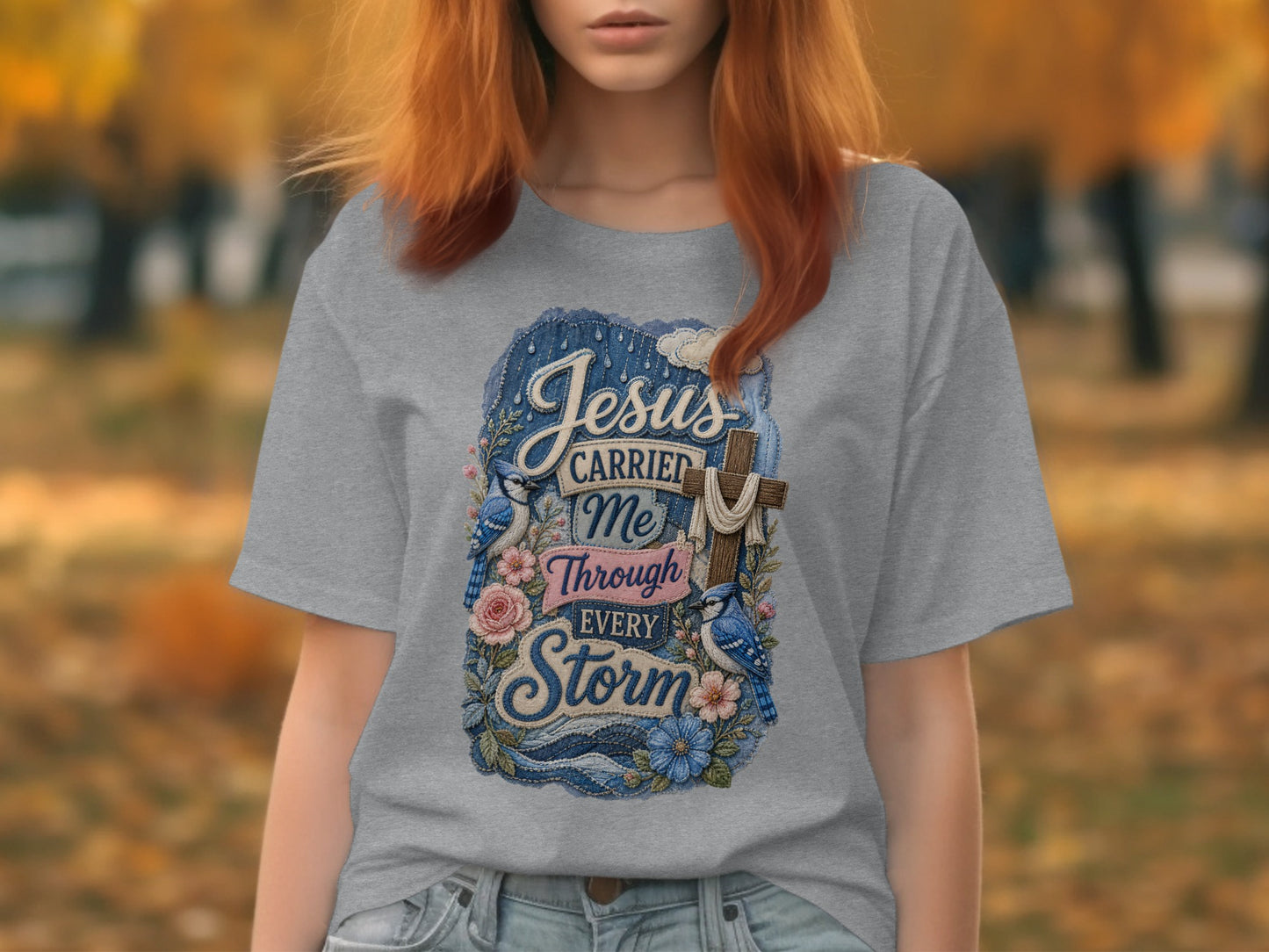 Gray t-shirt with blue distressed graphic of a cross, flowers, and birds with text "Jesus Carried Me Through Every Storm."