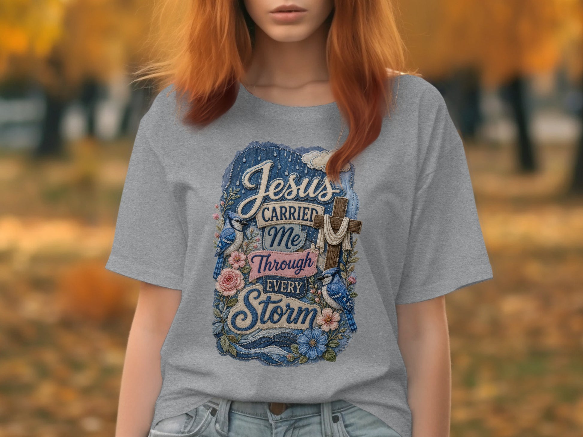 Gray t-shirt with blue distressed graphic of a cross, flowers, and birds with text "Jesus Carried Me Through Every Storm."