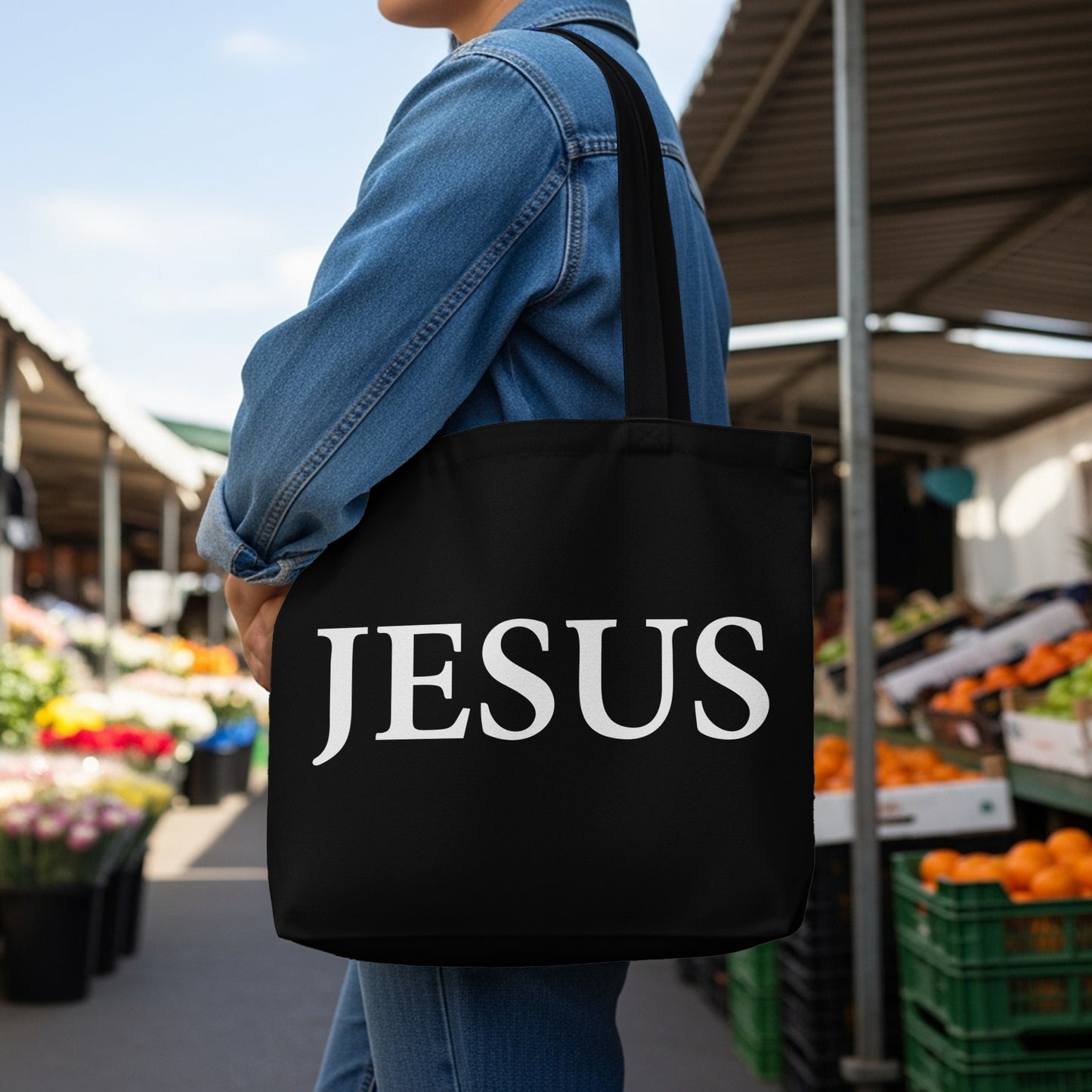 Black tote bag with "JESUS" printed in white letters, worn by a person in a denim jacket.