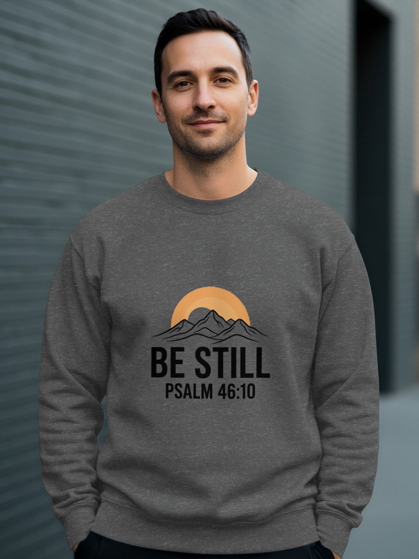 Man wearing gray sweatshirt with mountains, sun, and "BE STILL PSALM 46:10" text.