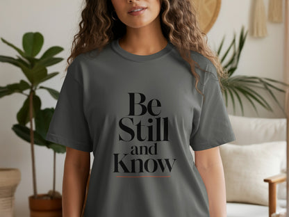 Dark grey t-shirt with black text "Be Still and Know" and an orange line.
