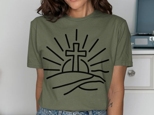 Olive green t-shirt with a black line drawing of a cross rising over hills at sunrise.