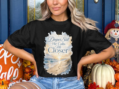 Black t-shirt with "Deeper Still, He Calls Me Closer" text over a beach scene.