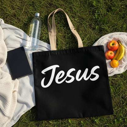 Black tote bag with white "Jesus" text on a picnic blanket with fruit and water.