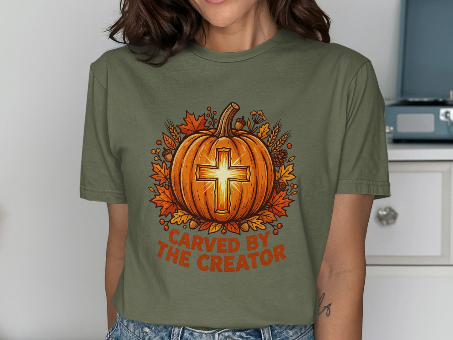 Olive green t-shirt with carved pumpkin and cross, surrounded by autumn leaves.