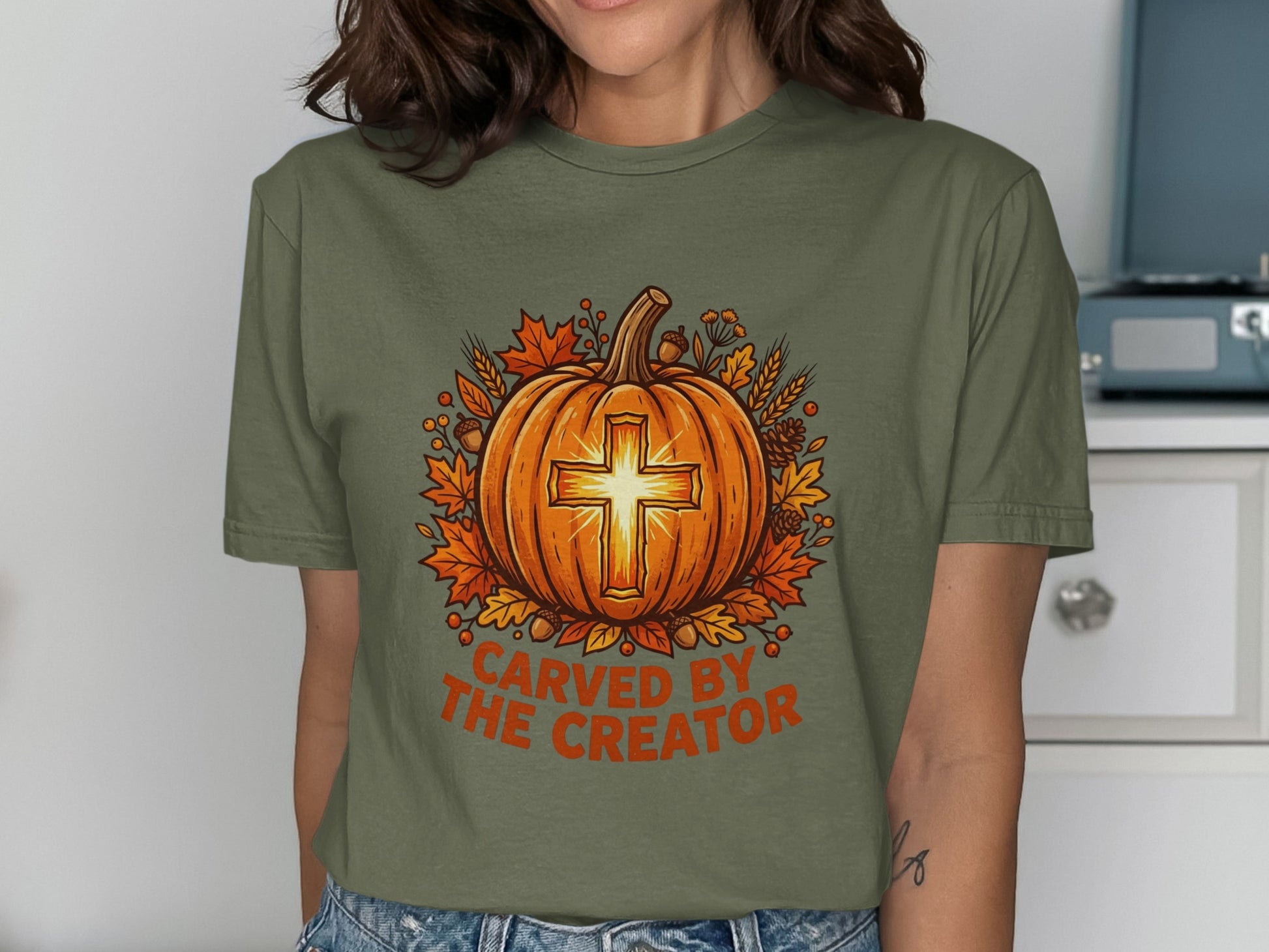 Olive green t-shirt with carved pumpkin and cross, surrounded by autumn leaves.