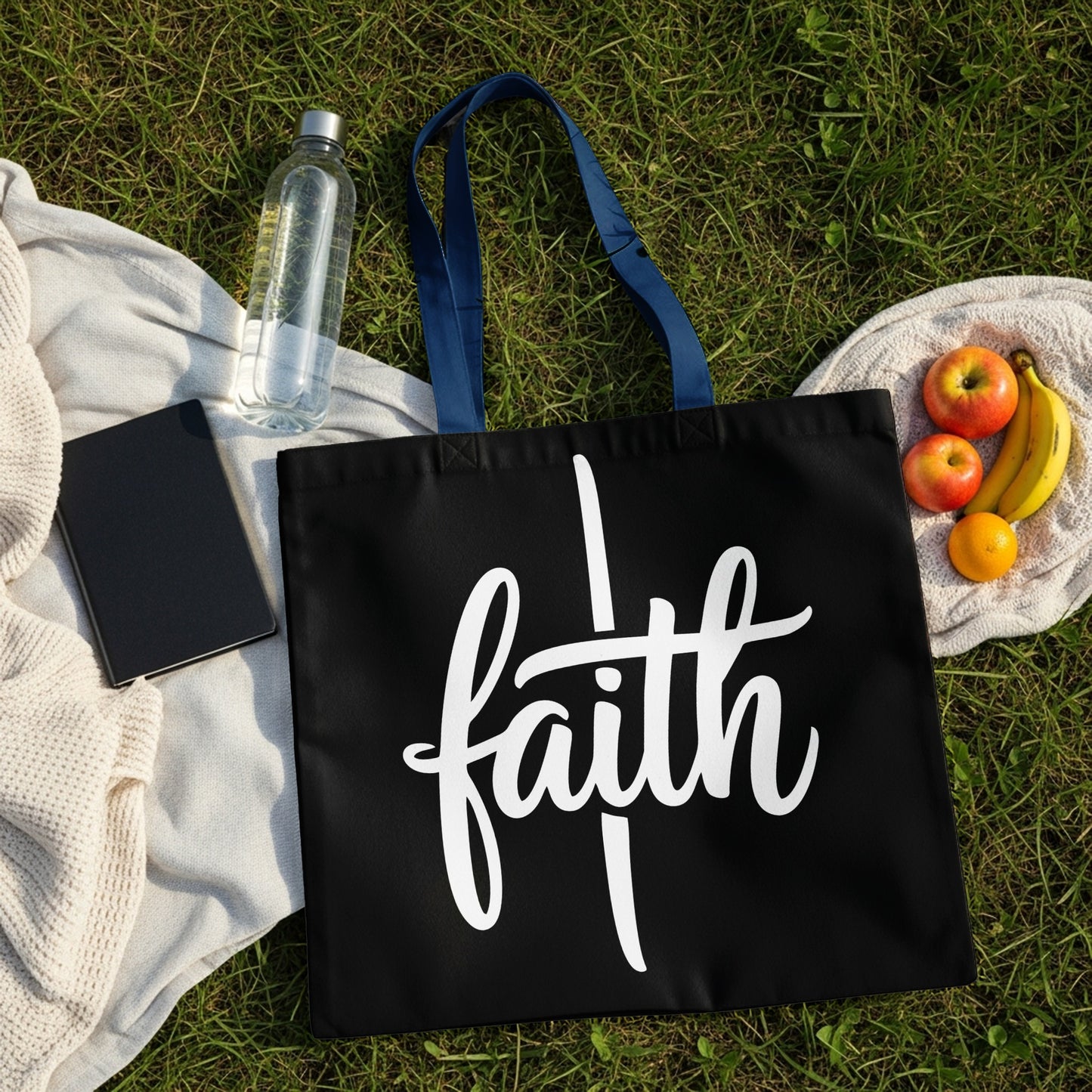 Black tote bag with white "faith" script, picnic items on grass.