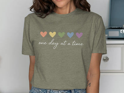 Olive green t-shirt with pastel hearts and "one day at a time" text.