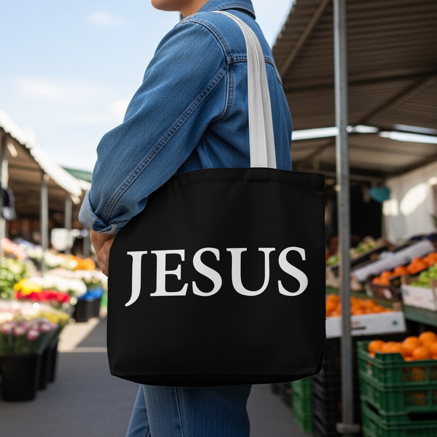 Black tote bag with white "JESUS" text worn by person at outdoor market.