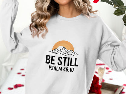 White sweatshirt with "Be Still Psalm 46:10" text and mountain sunset graphic.