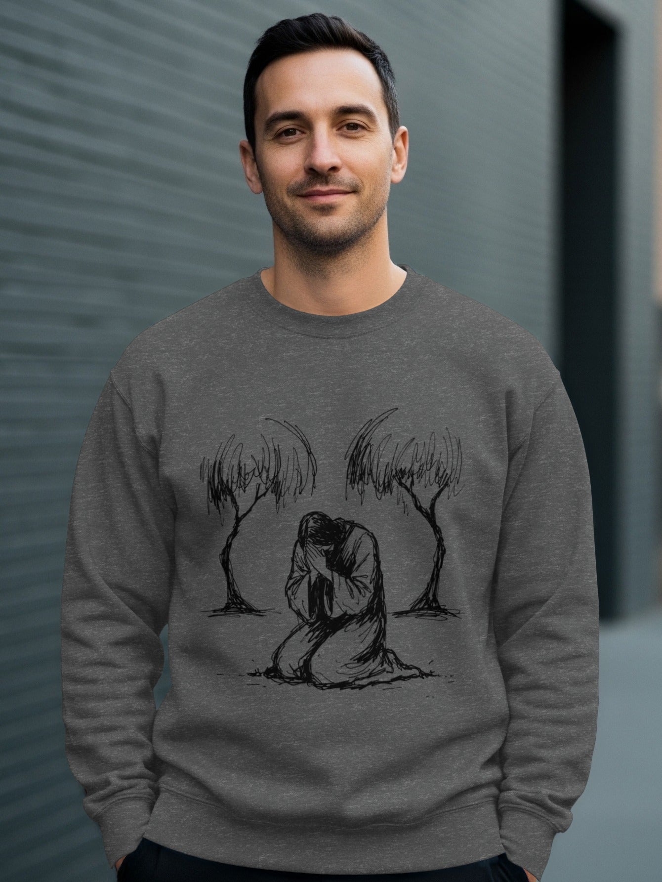Man wearing a grey crewneck sweatshirt with a sketch of a kneeling figure between two trees.