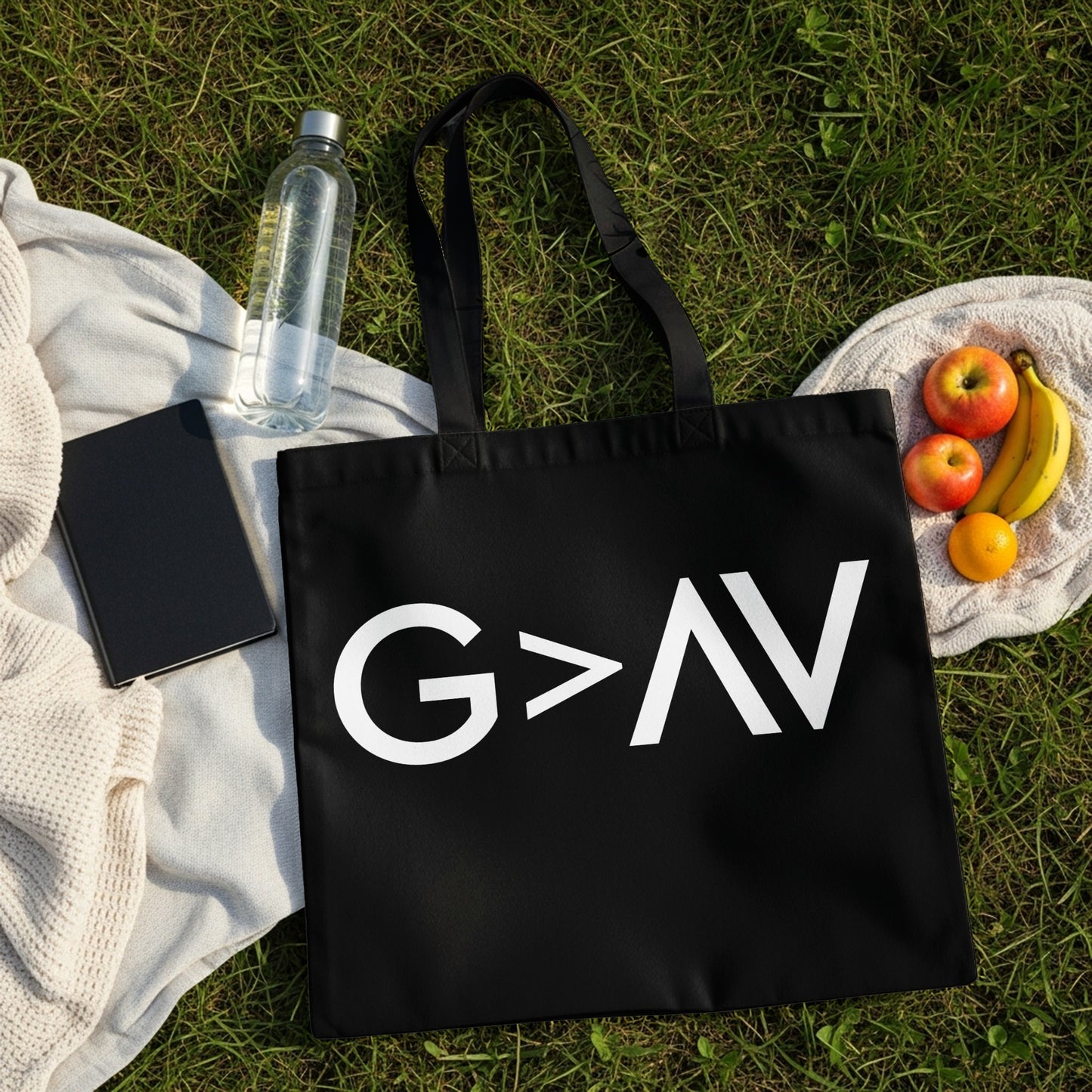 Black tote bag with white "G>AV" graphic, on grass with fruit and water bottle.