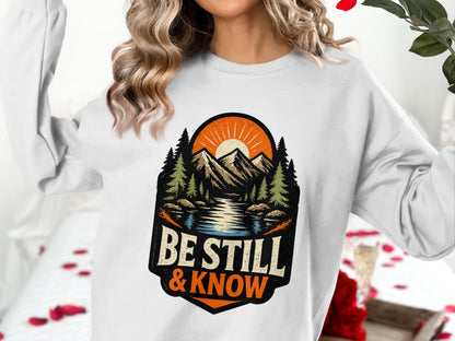 White sweatshirt with "Be Still & Know" text and mountain landscape graphic.
