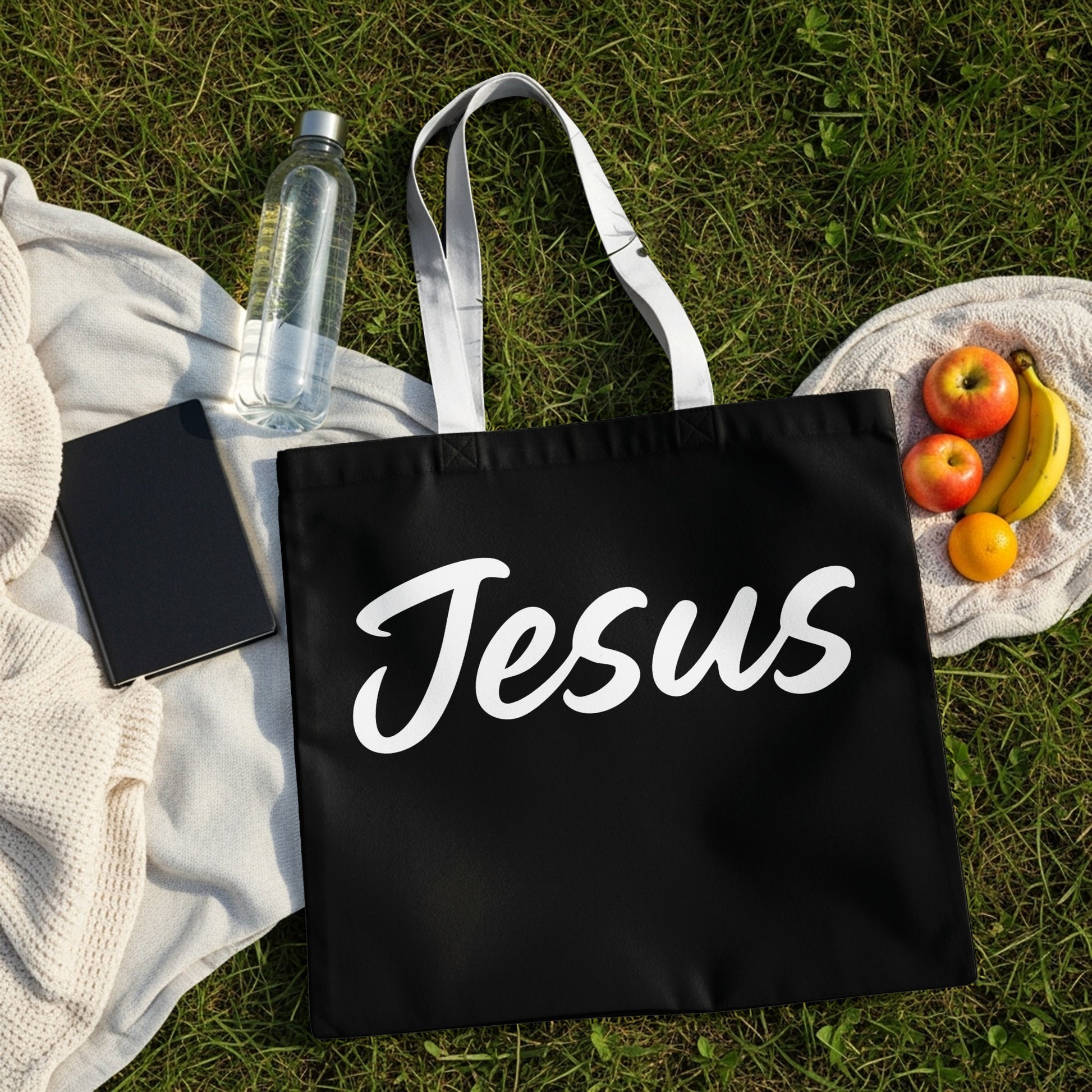 Black tote bag with "Jesus" in white script, on grass with fruit and water bottle.