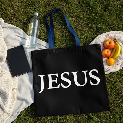 Black tote bag with "JESUS" printed in white, laid out on grass with fruit.